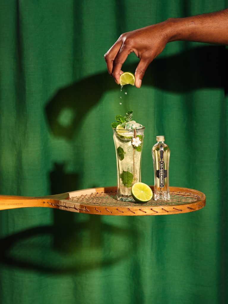 St-Germain bottle and lime cocktail on tennis racket with green backdrop, commercial drinks photography