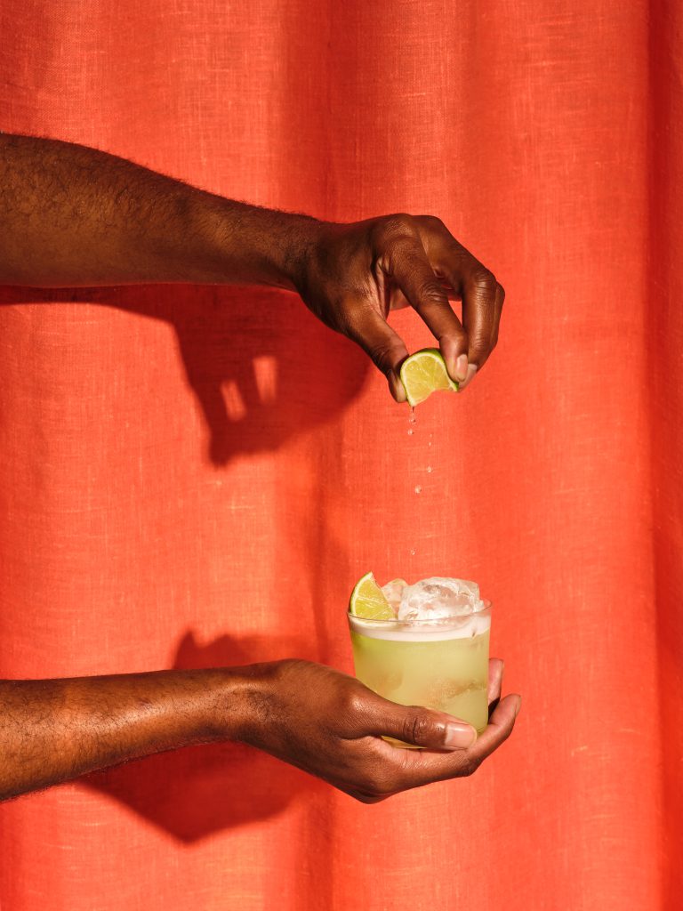 Hands squeezing citrus over St-Germain cocktail against red backdrop
