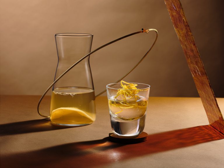 Cocktail with citrus garnish beside glass carafe on warm-toned set, editorial still life