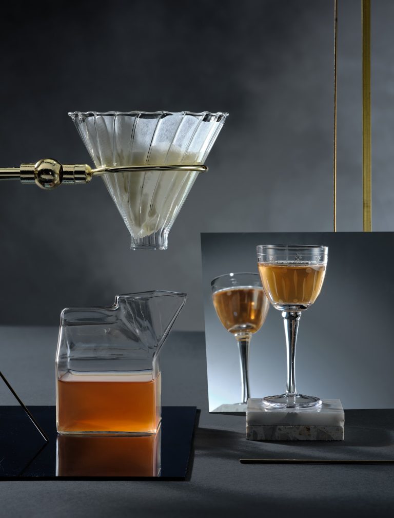 Glass funnel pouring liquid into carafe with cocktail glasses on geometric set