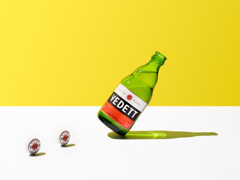 Vedett beer bottle tilted on white surface with yellow background, studio still life